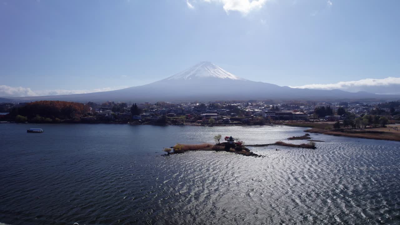 Plano aéreo en retroceso tomado con dron que muestra la grandiosidad del monte Fuji con el lago Kawaguchi y el paisaje urbano circundante bajo cielos despejados