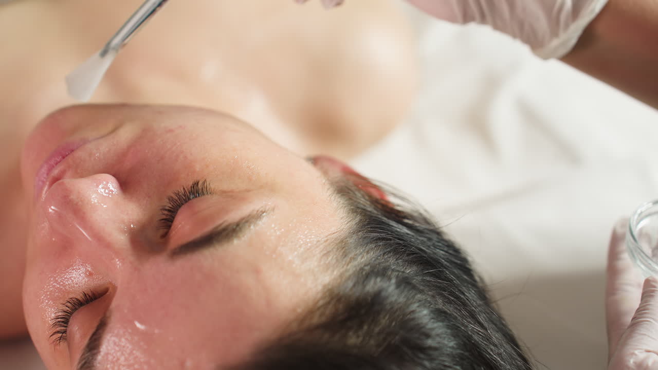 High angle of woman lying with eyes closed on spa bed while cosmetologist in gloves dips brush into gel and applies nourishing facial mask in gentle strokes for glowing radiant skin care