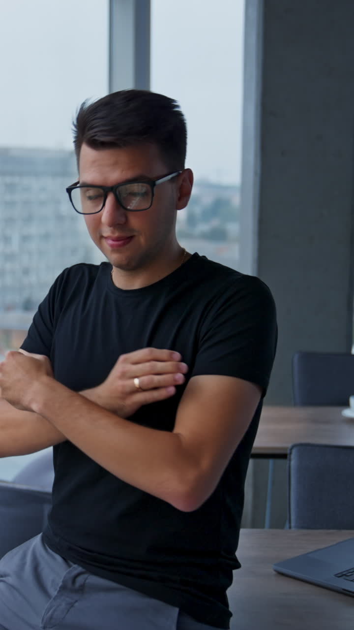 Smiling confident young businessman in the light office. Man puts on glasses looking into camera. Blurred backdrop. Vertical video