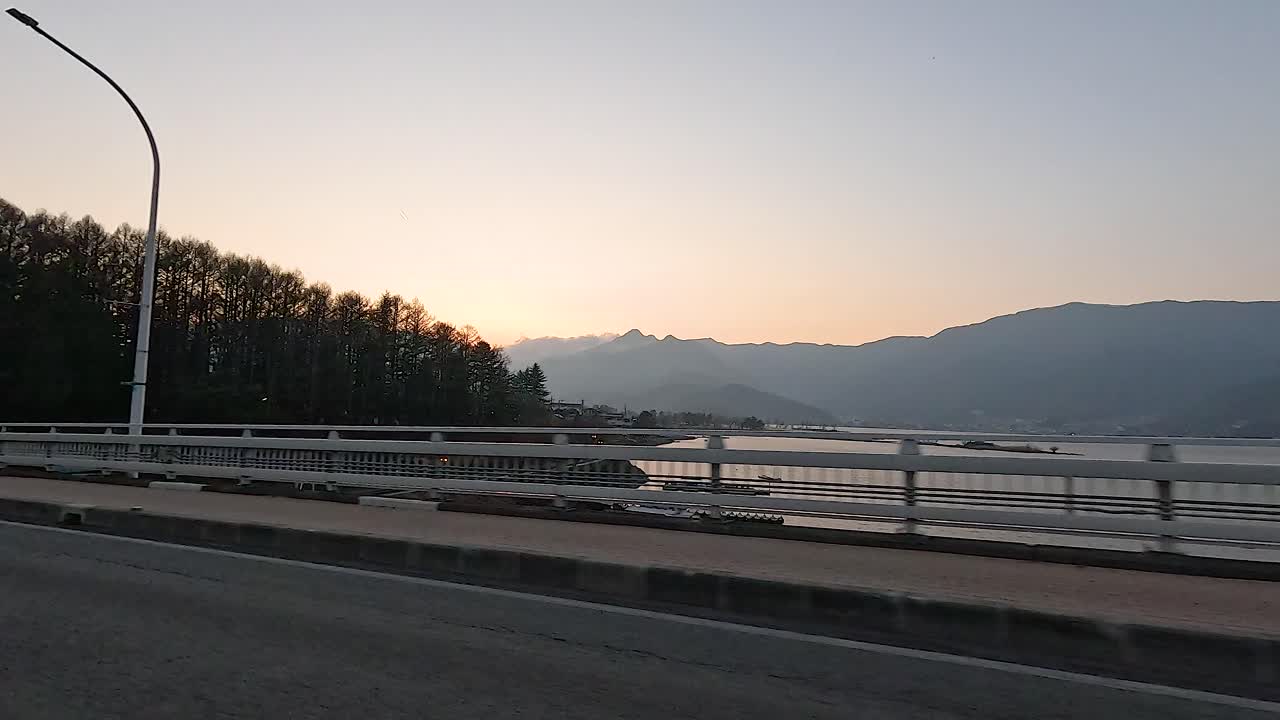 A serene morning drive captures the tranquil beauty of Lake Kawaguchi with silhouetted trees and distant mountains under soft dawn light