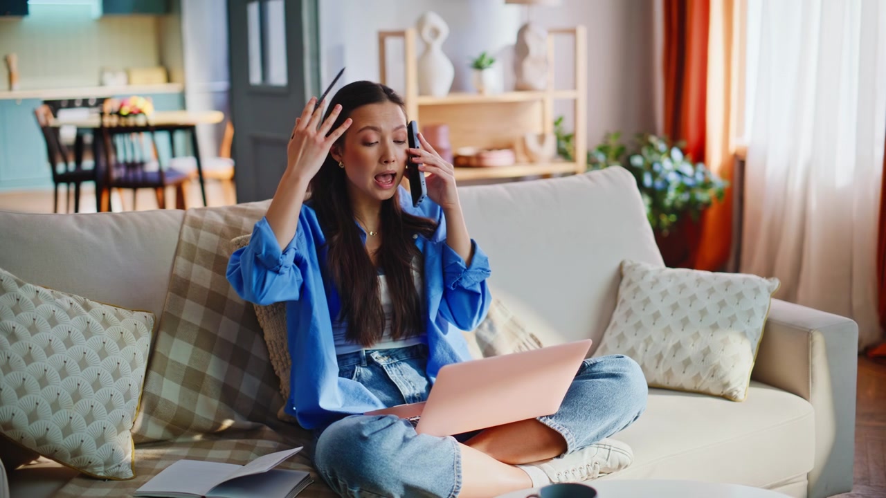 Asian freelancer gesturing phone call working pink laptop sofa. Remote multitasking