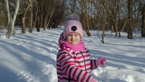 Child-girl-throwing-snowball-towards-camera,-smiling-kid-walking,-playing-with-snow-in-winter-park