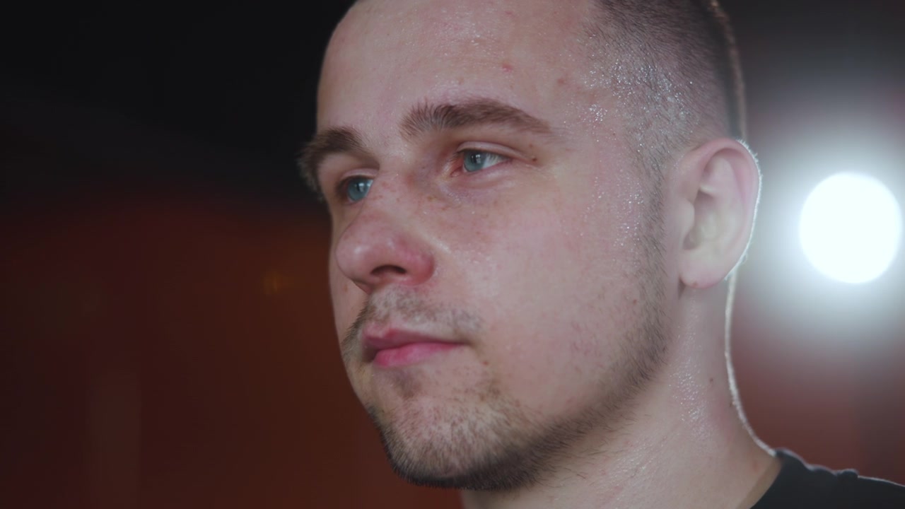 Primo piano ravvicinato della testa e della parte superiore del collo di un giovane adulto maschio in una calda luce interna, con sudorazione, barba corta, acne lieve e una respirazione sottile che suggerisce un recupero post-allenamento, ritratto maschile.