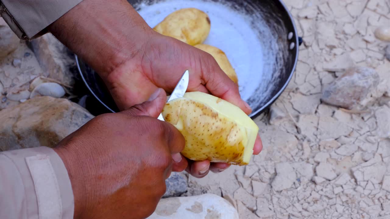 primer plano de las manos de un hombre pelando papas con una navaja de bolsillo en un entorno natural al aire libre preparando el almuerzo en el campamento durante una caminata