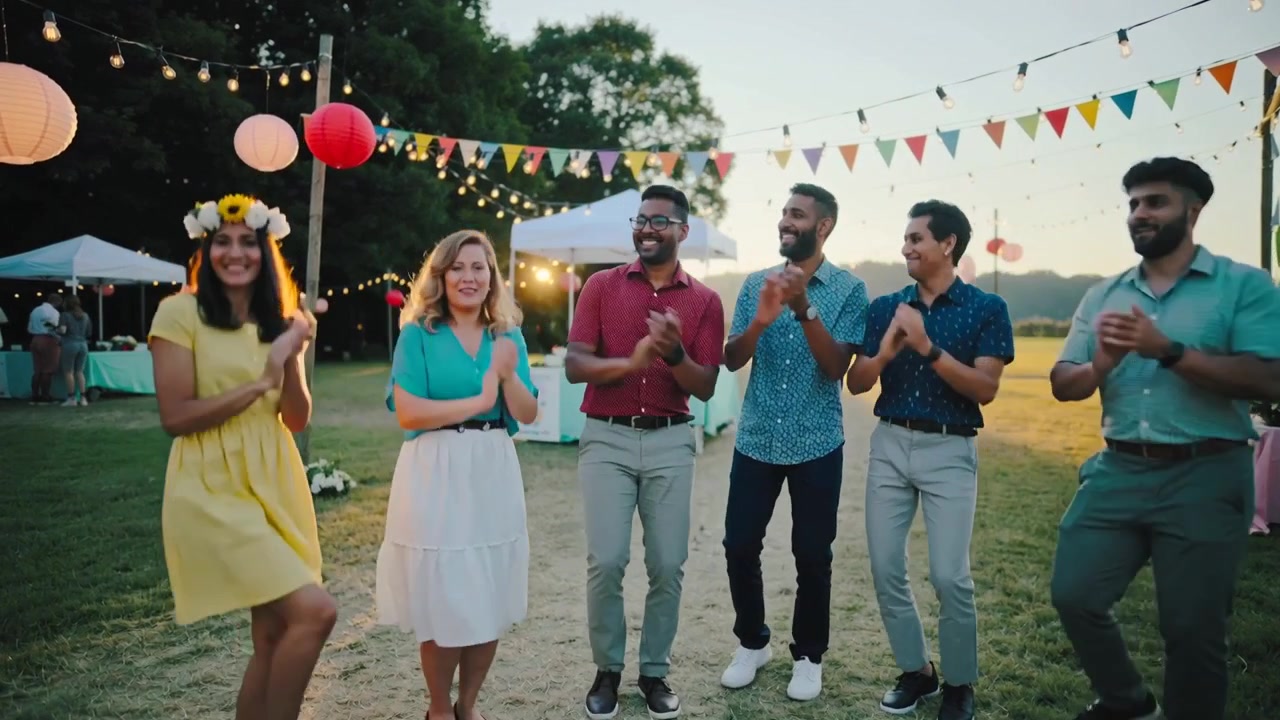 Six amis applaudissent et célèbrent en extérieur au coucher du soleil, avec une robe jaune, une couronne de fleurs et des guirlandes lumineuses.