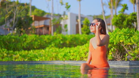 Fit-young-woman-relaxing-inside-the-outdoor-swimming-pool,-exotic-spa-hotel-wearing-an-orange-bikini-on-lush-greenery-background,-girl-touching-the-frame-of-sunglasses