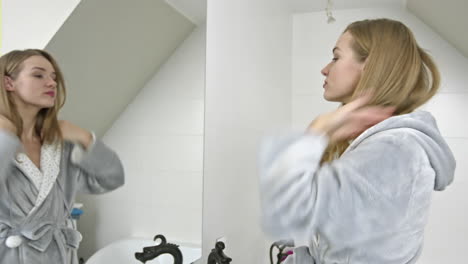 beautiful attractive young woman wearing bathrobe at bathroom standing next to mirror and checking her skin on face and hairs