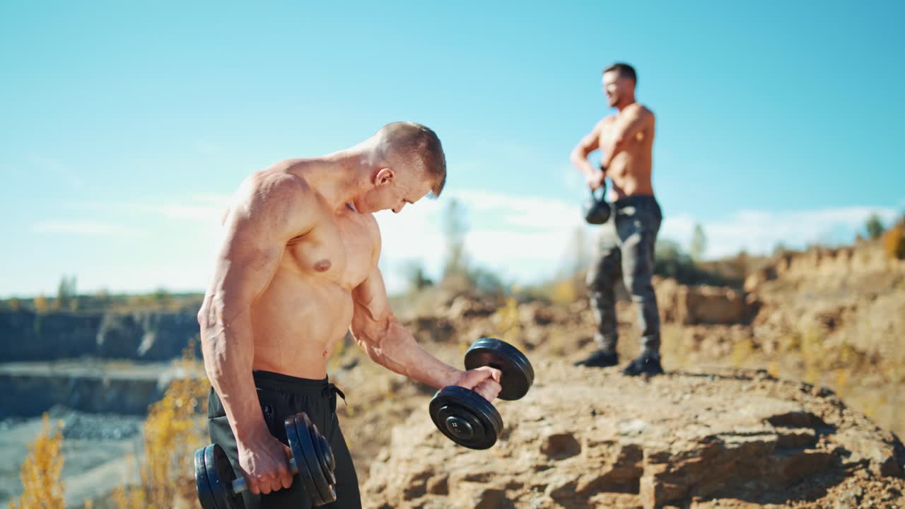Sportsmen doing workout outdoors. Muscular athlete is lifting dumbbells in hands on the background of a shirtless man with kettlebell in summer.