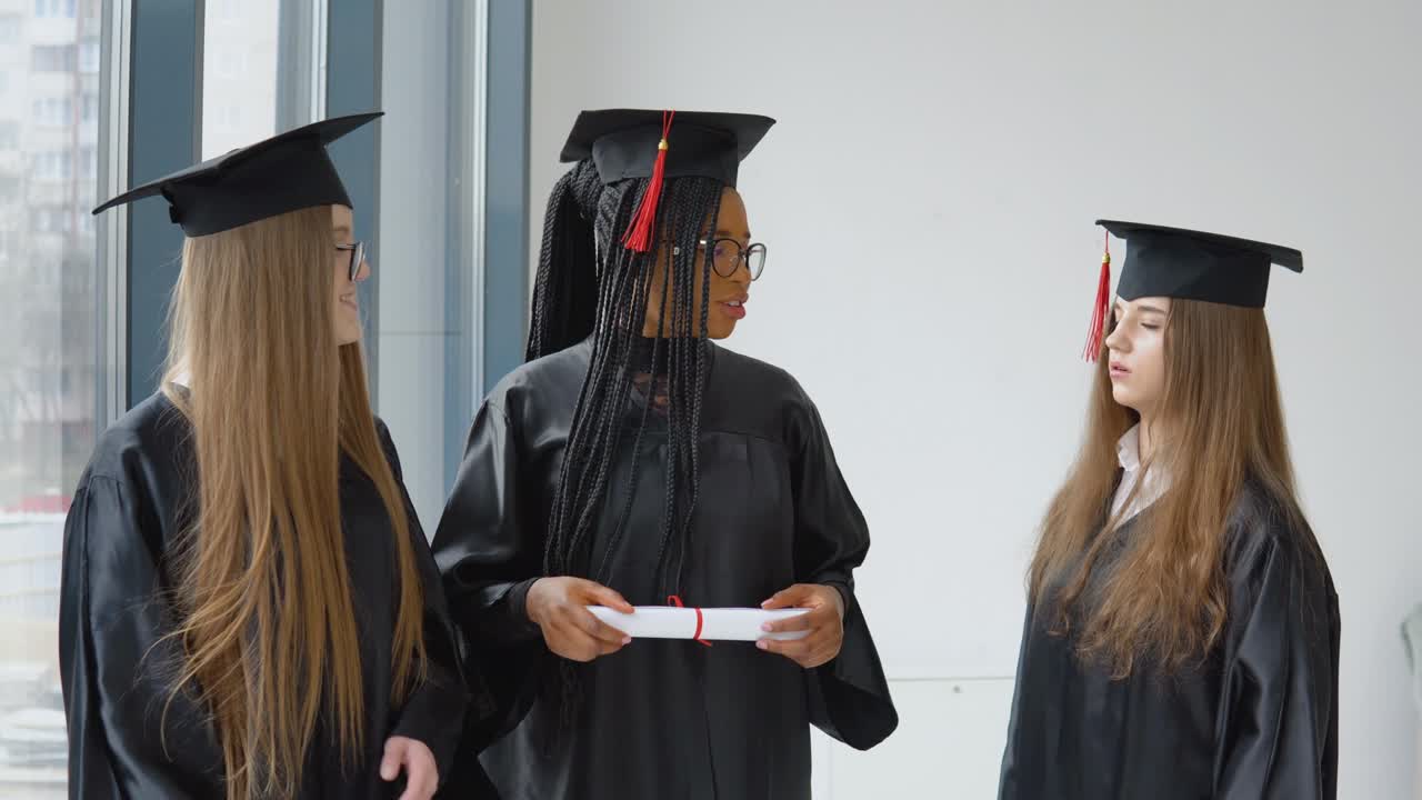 tres estudiantes caucásicas y afroamericanas con un diploma en sus manos. graduados con túnicas negras y sombreros cuadrados se comunican juntos. concepto de educación