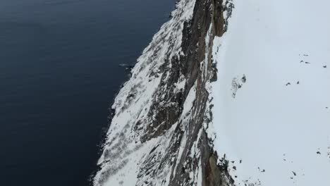Drone-view-in-Tromso-area-in-winter-flying-over-a-snowy-mountain-and-looking-down-the-cliff-into-the-fjord-in-Swgla,-Norway-top-view