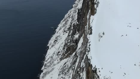 drone view in tromso area in winter flying over a snowy mountain and looking down the cliff into the fjord in swgla, norway top view