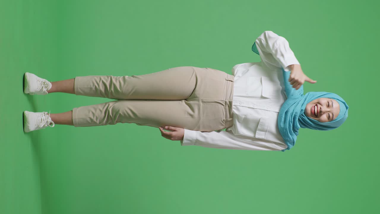 녹색 스크린 배경 스튜디오에 서있는 동안 카메라에 엄지손가락을 보여주고 미소 짓는 아시아 무슬림 여성의 몸이 가득 차 있습니다.