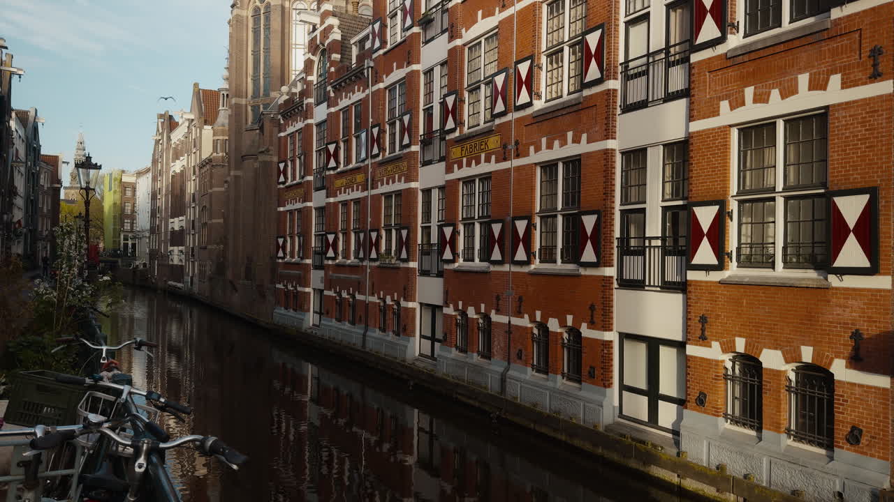 Klassisk Amsterdam-kanal omkranset af traditionelle murstenshuse med karakteristiske røde og hvide skodder, med spejlbilleder i vandet og cykler i forgrunden.