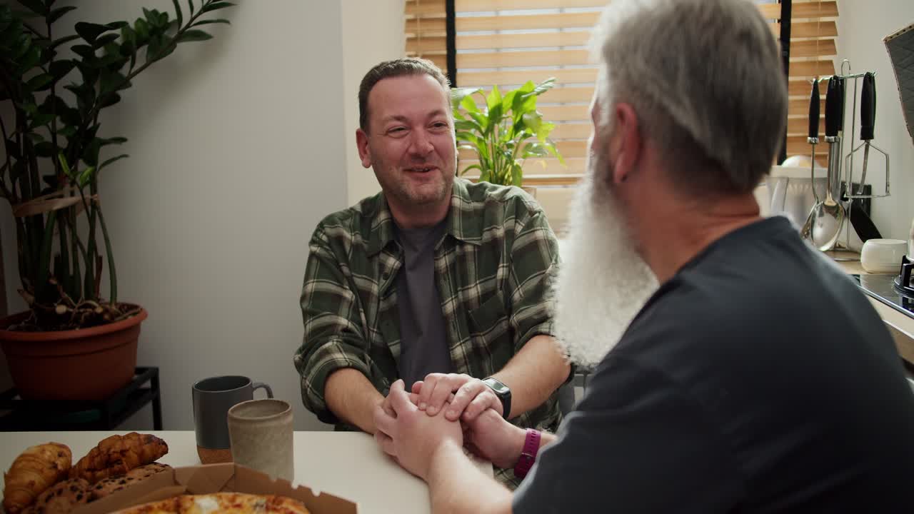 Over the shoulder a happy brunette man with stubble in a green plaid shirt holds hands with his older boyfriend with gray hair and a lush gray beard in a gray T-shirt during his conversation in the kitchen at the table