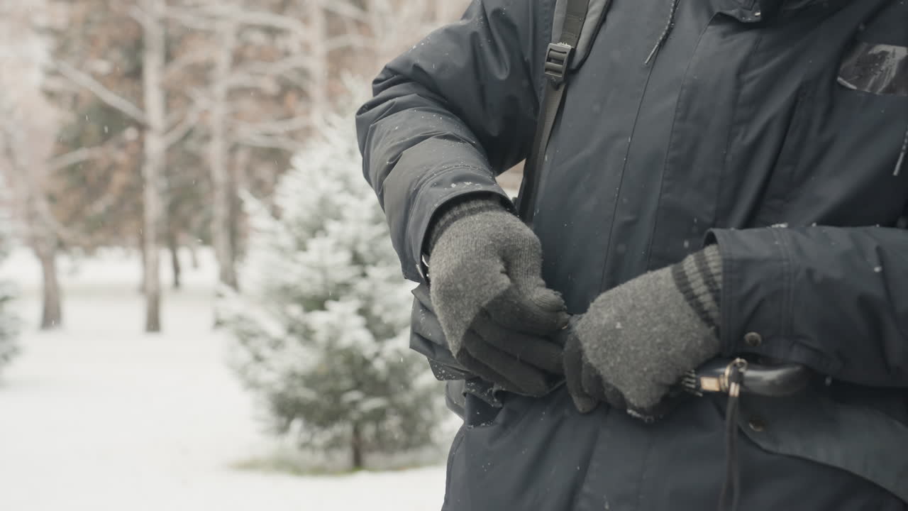 Pria mengaitkan karabiner ke tali jaket di taman bersalju, sarung tangan rajut dan mantel abu-abu, close-up tangan mengaitkan klip logam dan menyesuaikan gesper, pohon cemara kecil dan salju yang turun menciptakan suasana perkotaan yang tenang.