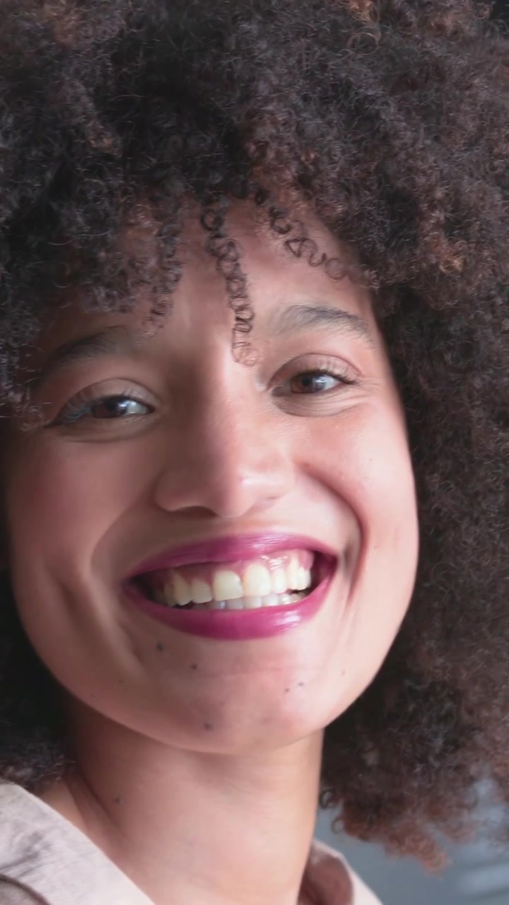 Vídeo vertical: Mulher abrindo os olhos, sorrindo e rindo para a câmera em estúdio com ripas, vestindo camisa bege.