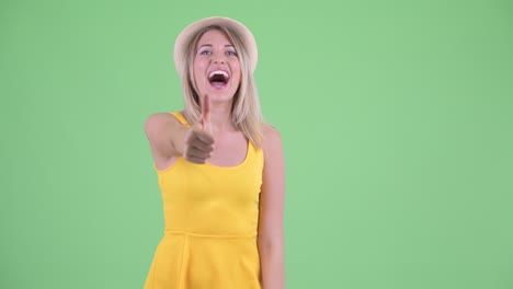 studio shot of young tourist woman looking excited and giving thumbs up against green background