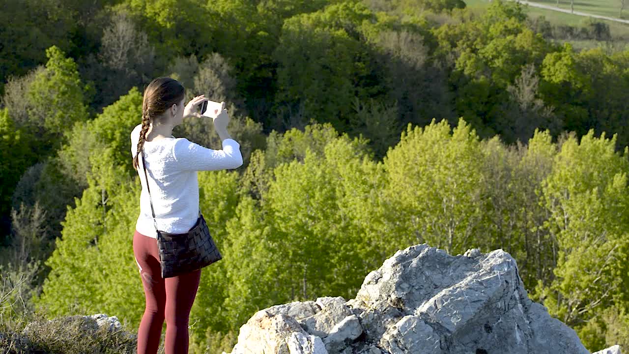 chica tomando fotos de la naturaleza con su teléfono móvil mientras está de pie en la cima de la colina cerca de enormes rocas