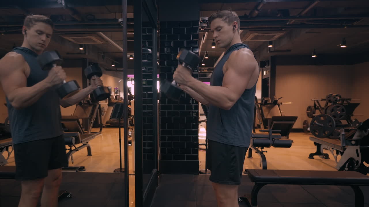 Fit Man Lifting in Front Of Mirror Doing Hammer Bicep Curls Weight Exercise - Blond Athletic Sportsman Training Forearm Muscle Flexing and Pumping Arms While Listening Music in Gym