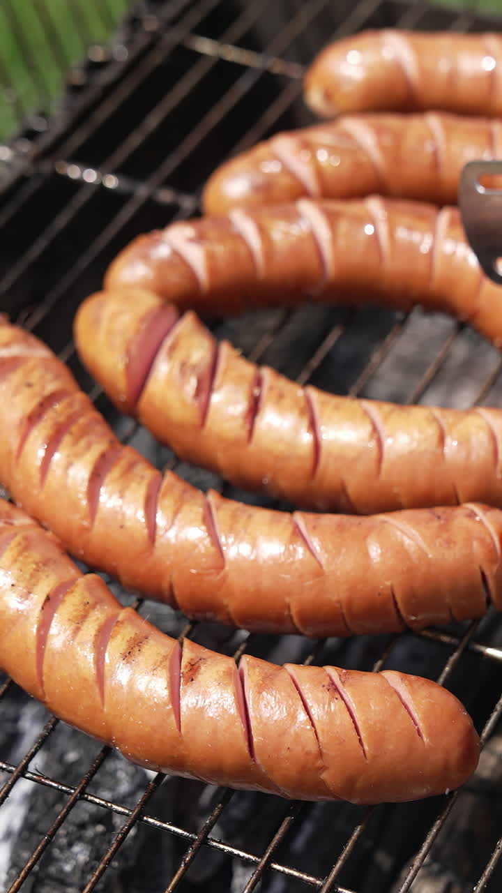 Tasty juicy sausages grilling. Barbecue grill cooking outdoors. Hot dog sausages are turning with tongs on a grill plate. Food for picnic. Vertical video