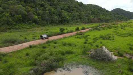 Camión-Moviéndose-En-Un-Camino-De-Tierra-Húmeda-En-El-Paisaje-Rural-De-América-Del-Sur-A-Lo-Largo-De-La-Selva-Y-Las-Marismas,-Disparo-De-Drones