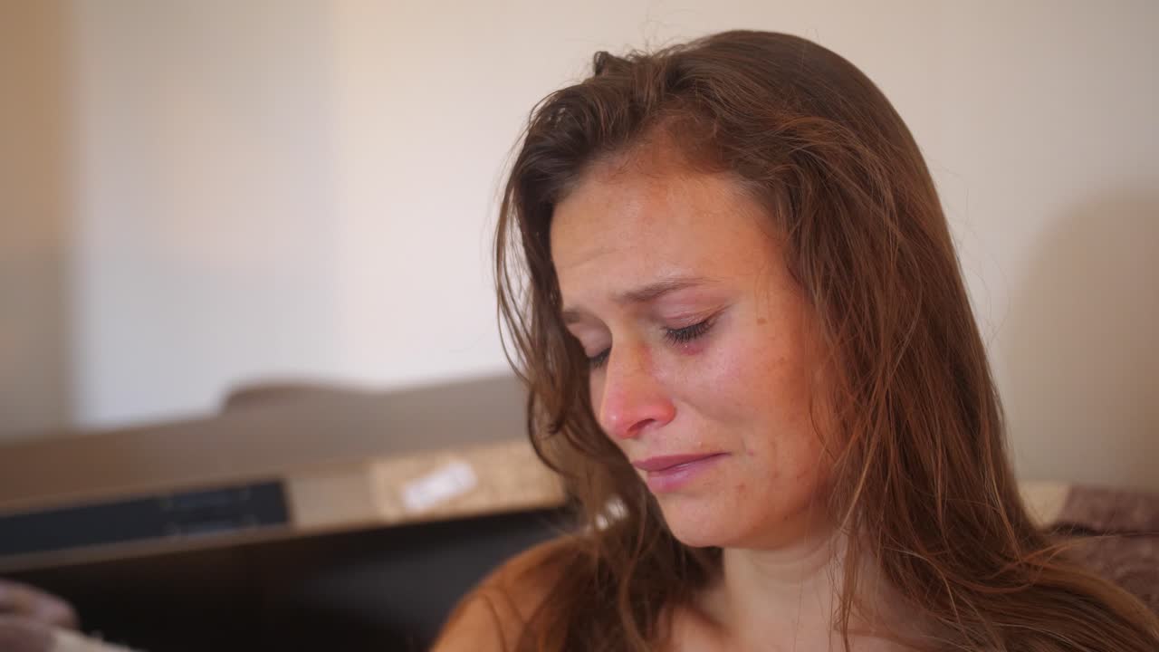 A close up portrait shot of a young woman with wounds and bruises, stares with a blank look and starts crying while touching her sore cheek