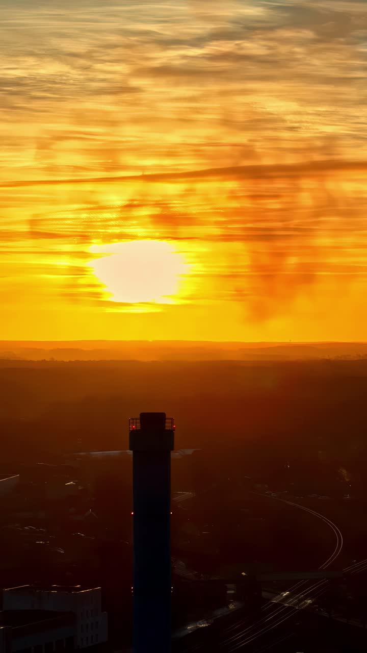 Industrial chimney silhouette against bright orange sunset, aerial vertical view