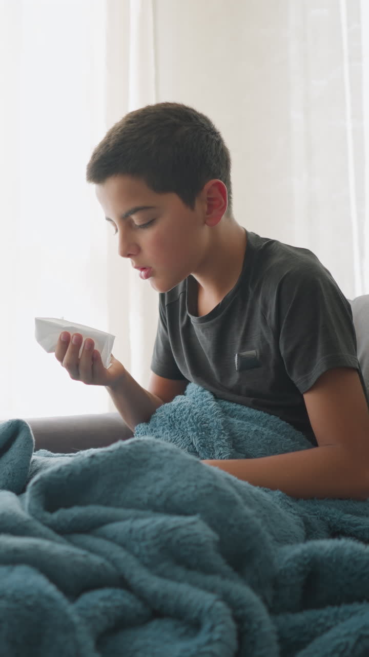 Young kid sneezing into tissue seated on couch under blue blanket, with tissue roll nearby, displaying signs of illness, fatigue, and recovery, showing home care in the living room