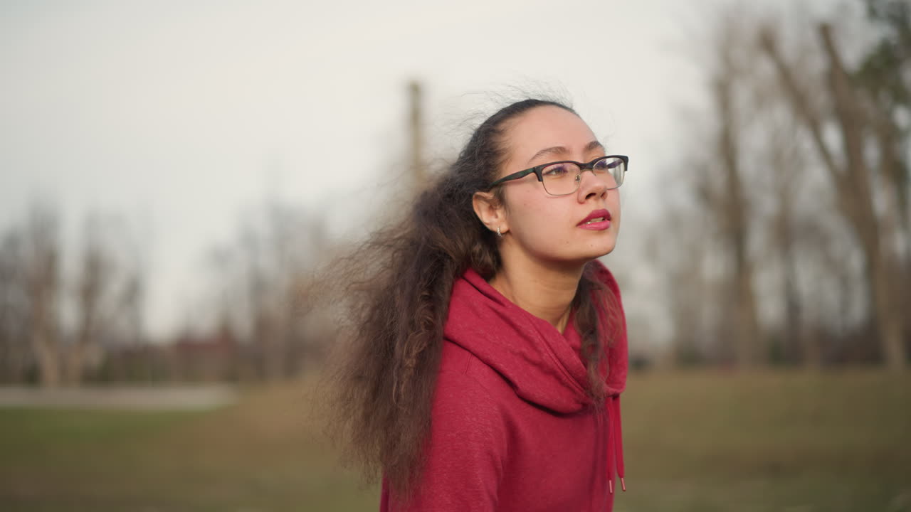 Asian Woman Standing In Park CloseUp Portrait, Calm Expression With Soft Gaze, WindBlown Hair And Glasses, Hoodie Collar Framing Face, Muted Winter Trees In Background Create Introspective Mood