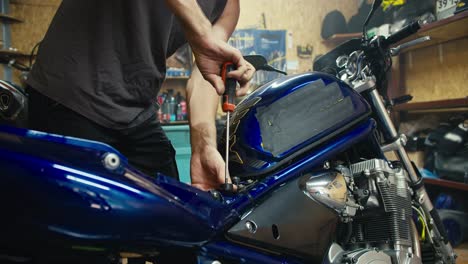 Primer-Plano:-Un-Hombre-Con-Una-Camiseta-Gris,-Un-Mecánico-Con-Una-Camiseta-Gris,-Repara-Una-Motocicleta-Azul-Con-Un-Destornillador-En-El-Taller-De-Su-Garaje.