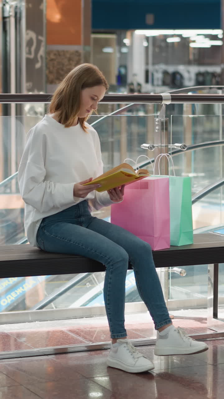 joven sentada en un moderno centro comercial leyendo un libro amarillo mientras balancea su pierna ligeramente, rodeada de bolsas de compras coloridas, barandillas de vidrio, plantas en maceta, escaleras mecánicas