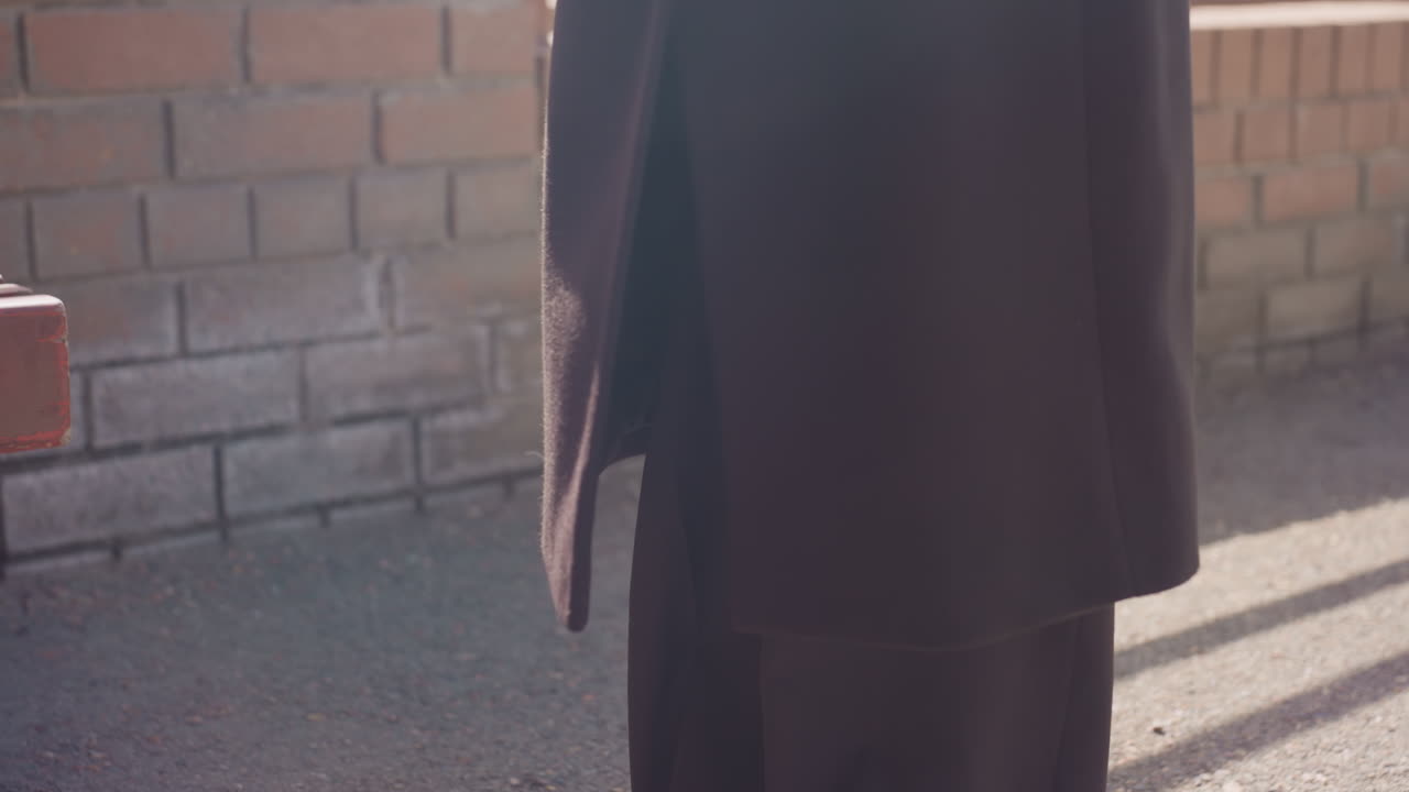 검은색 코트를 입고 검은색 바지를 입은 사람이 산들바람에 흩날리는 모습이 아래에서 보이는 구도예요. 햇빛이 벽돌 벽 근처에 그림자를 드리우고, 옷감은 부드럽게 움직이며 스타일과 시원한 아침 공기가 느껴지는 도시의 산책로 풍경입니다.