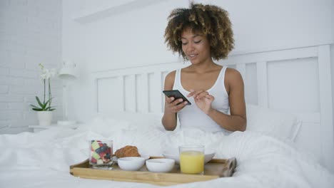 Woman-with-smartphone-in-bed-having-meal
