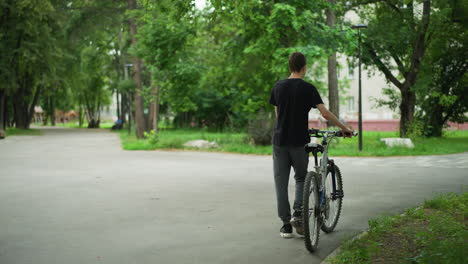 young boy in black top and ash pants walking alongside his bicycle in park pathway, trees and greenery line the path, while distant buildings and more trees are visible