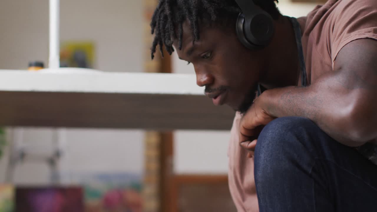 Close-up de artista masculino afro-americano usando fones de ouvido, pintando em tela no estúdio de arte.