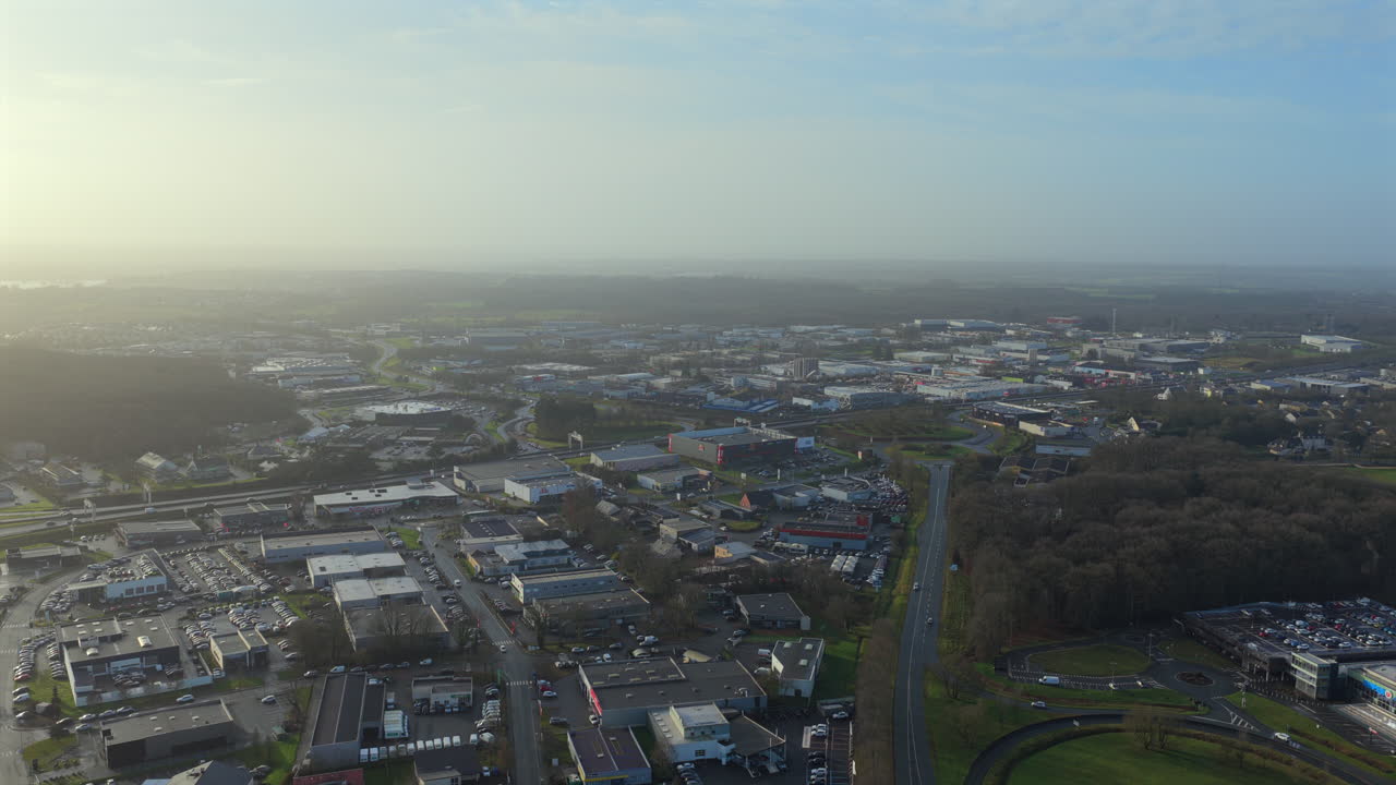 Aerial drone glide over an industrial and commercial zone near Angers, Maine et Loire, France, showing warehouses, factories, roads, roundabout, green areas, and suburban landscape under soft daylight
