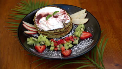 Primer-Plano-En-Cámara-Lenta-Del-Plato-Con-Saludables-Panqueques-Verdes-De-Avena-Y-Matcha-Servidos-Con-Rodajas-De-Manzana,-Kiwi-Y-Fresa-Y-Crema-Batida