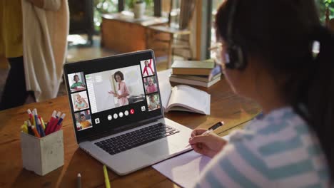 Schoolgirl-using-laptop-for-online-lesson-at-home,-with-diverse-teacher-and-class-on-screen