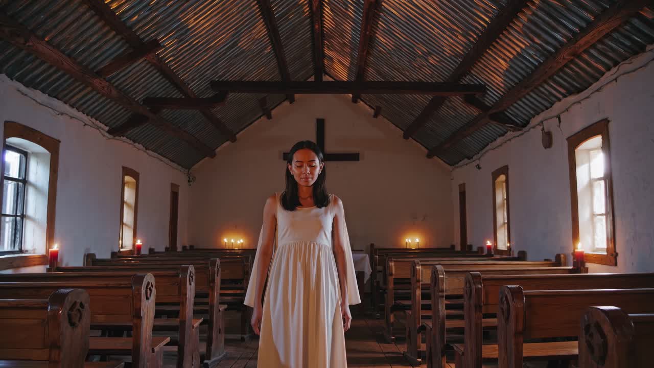 Young woman standing in a simple church with lit candles, experiencing a moment of devotion and tranquility under a metal roof and wooden cross