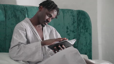 man in white bathrobe sitting on bed with green velvet headboard, focused on reading black notebook, turning pages, relaxed, cozy indoor atmosphere, soft lighting