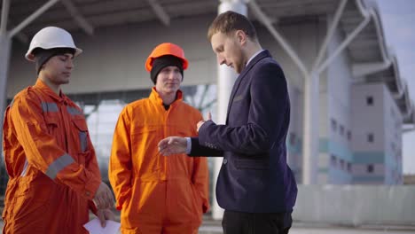 Manager-in-a-suit-giving-envelopes-with-money-to-workers-in-orange-uniform-and-helmets.-Successful-finish-of-the-project.-Shot-in-4K