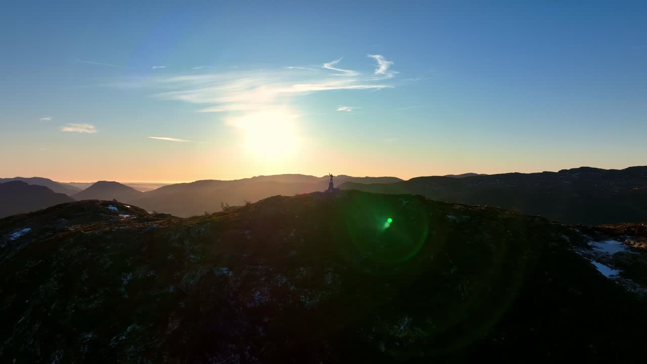 Silhouette of a person on a mountain peak raising an arm in victory as drone orbits at sunset