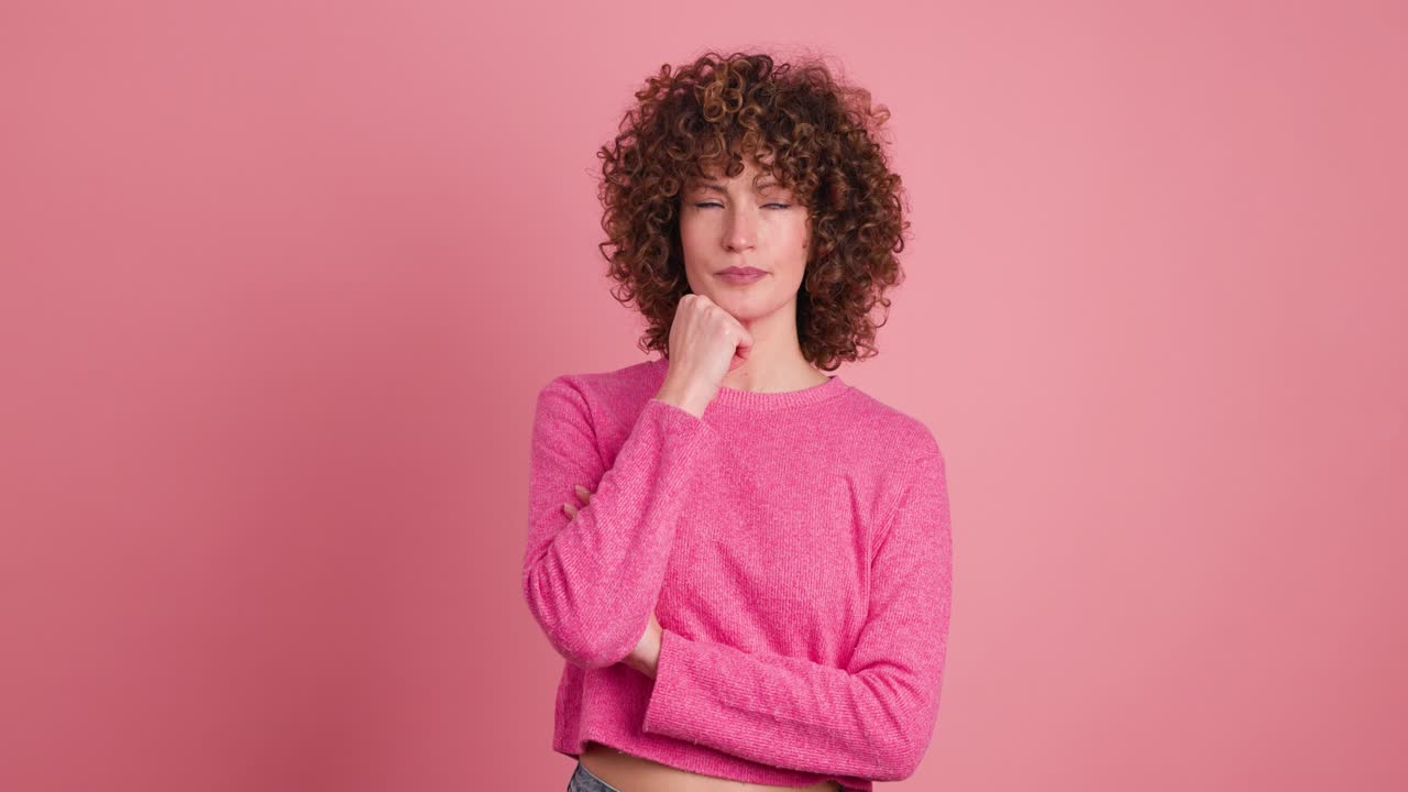 Thoughtful young woman touching face and smiling on pink background