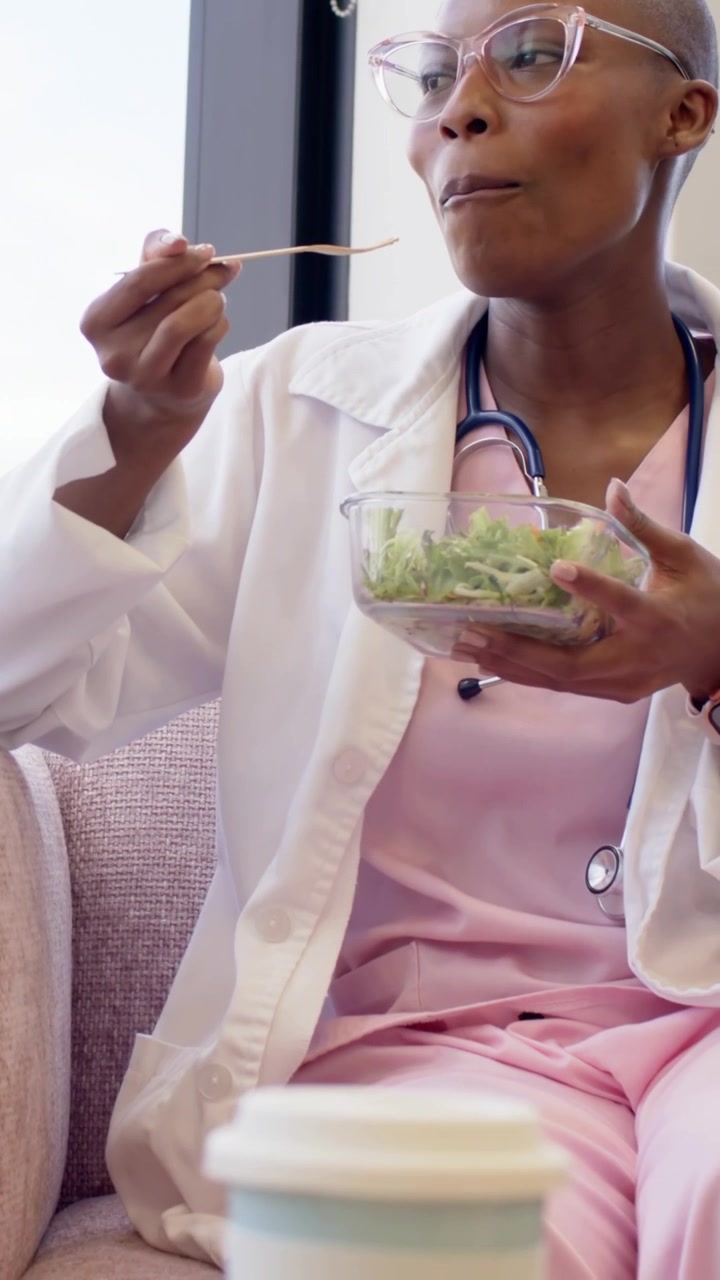 Video verticale: Infermiera in pausa, in divisa rosa e camice, mangia insalata con la forchetta, tazza in sala relax.