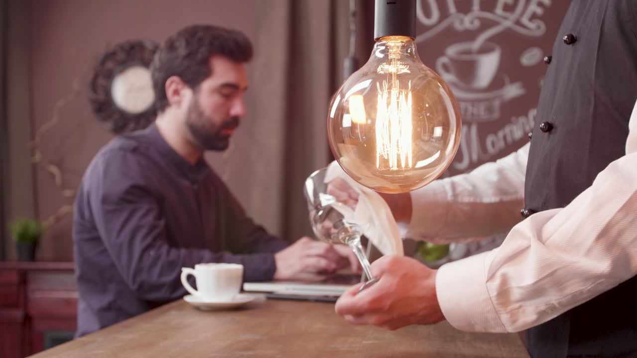 A man works on his laptop in a cafe while a waiter prepares a glass