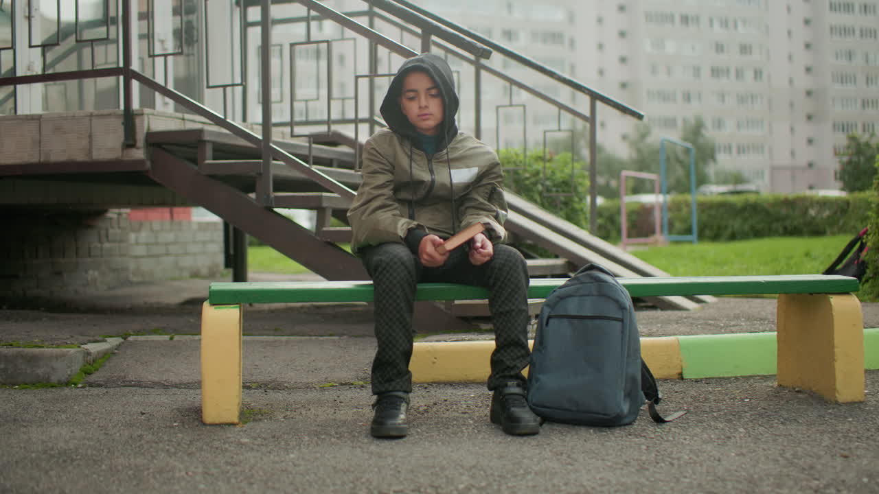 Student wearing hooded jacket seated on outdoor bench holding and tapping book on hand with backpack placed close by, staircase and residential buildings in background