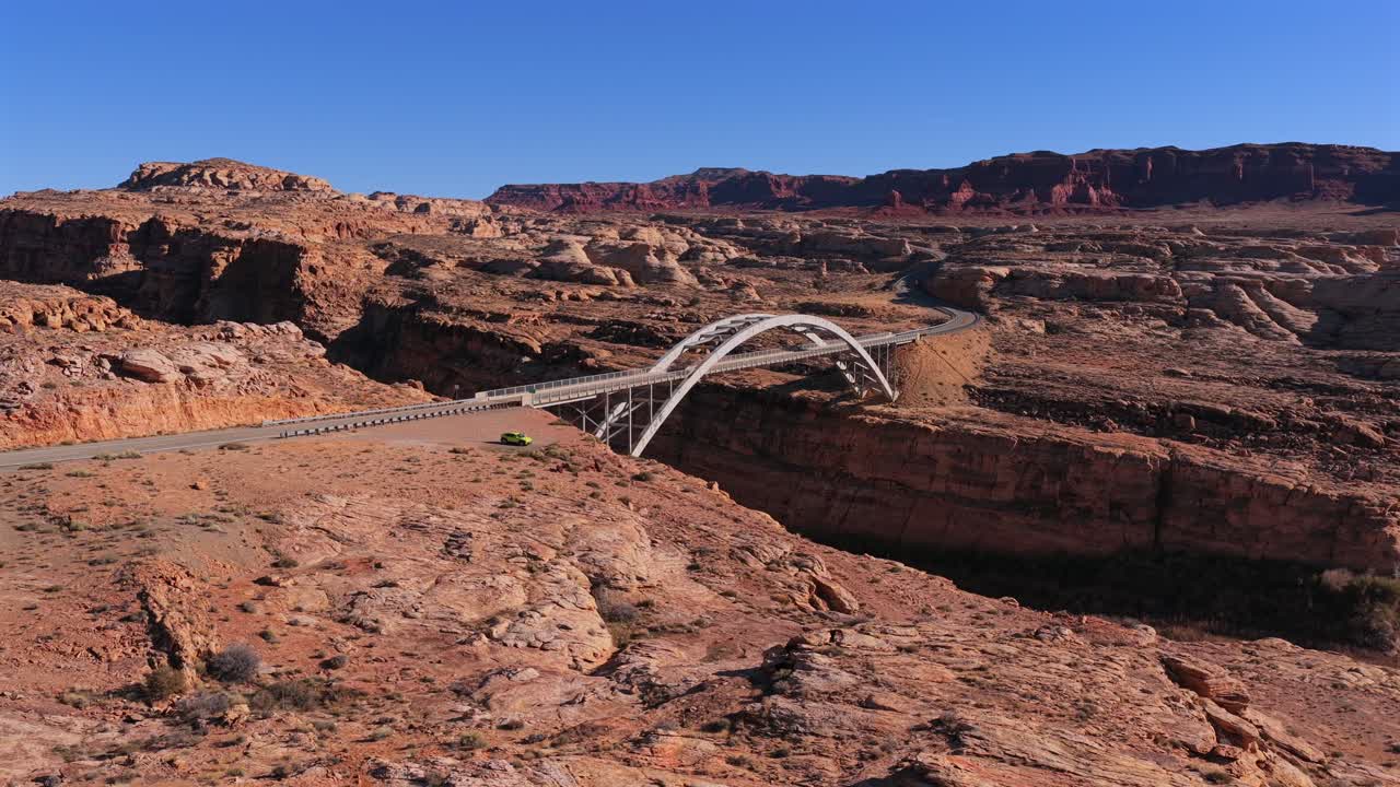 Una ripresa cinematografica con drone dall'alto cattura un elegante ponte ad arco che attraversa un ampio fiume. Scogliere rocciose rosse e aspre e profondi canyon desertici offrono uno sfondo mozzafiato per questa infrastruttura iconica.