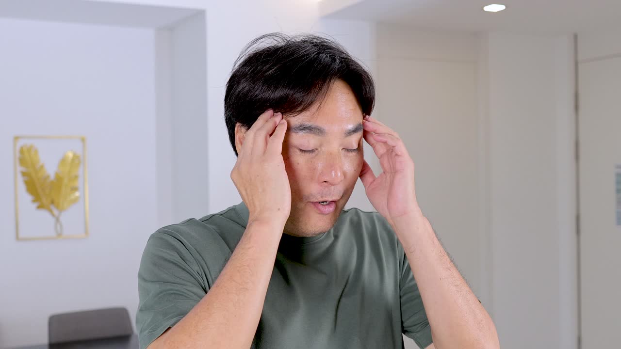 Asian man holds head with both hands, showing discomfort and stress in bright indoor setting