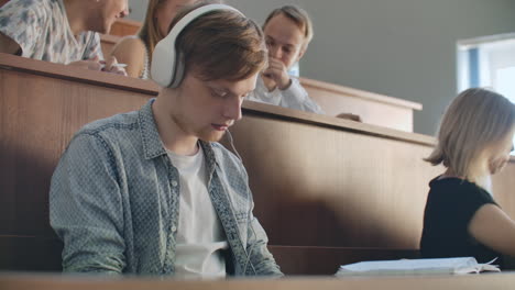 un estudiante masculino en un auditorio de la universidad escucha música con auriculares blancos durante un descanso sin prestar atención a los demás. mucha gente en la audiencia está hablando.