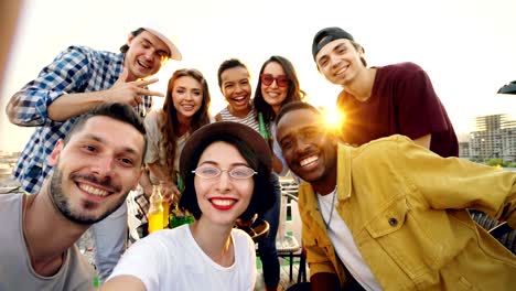 punto de vista de una mujer bonita con un sombrero de moda sosteniendo la cámara y tomando selfies con amigos disfrutando de una fiesta en la azotea con refrescos, la gente está mirando a la cámara y riendo.